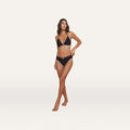 A woman stands barefoot against a white background, wearing the Miya Bikini Bottom featuring a ruched waistband and gold coin detail. She has shoulder-length brown hair, gazes slightly to the side, and rests her left hand on her thigh.
