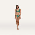 A woman stands against a plain white background wearing the Miya Bikini Bottom, featuring a green butterfly floral print and a sash waistband, looking toward the camera.