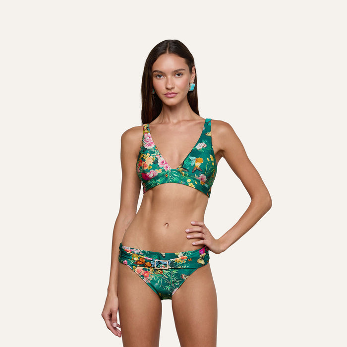 A woman stands against a plain light background with one hand on her hip, wearing the Miya Bikini Bottom featuring a green butterfly floral print and a sash waistband.