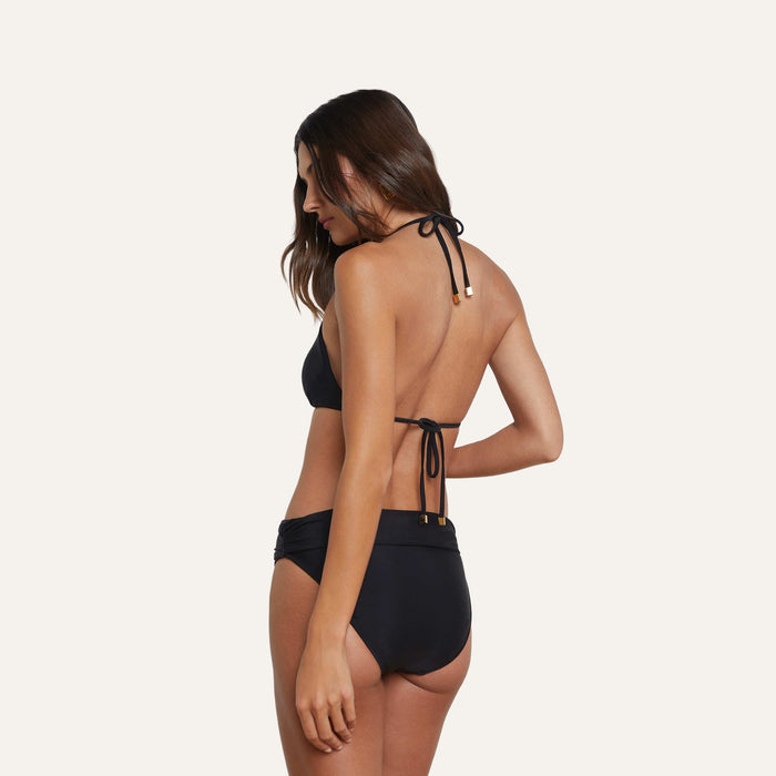 A woman with long brown hair, wearing the Miya Bikini Bottom featuring a ruched waistband in black, stands turned away from the camera, displaying the back of her swimsuit against a plain, light-colored background.