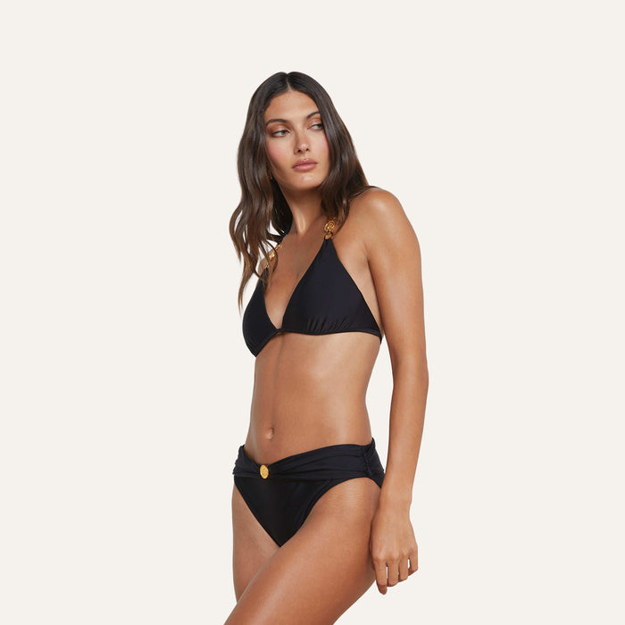 Model wears the Miya Bikini Bottom, featuring a black design with gold coin detail and a ruched waistband, standing against a plain light background and gazing to her left with a relaxed expression.