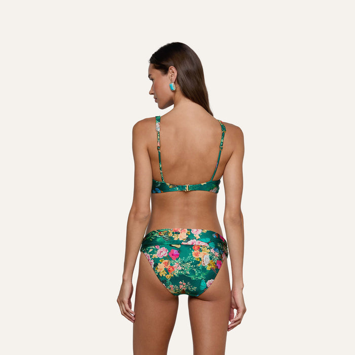A woman with long brown hair, wearing the Miya Bikini Bottom in a green butterfly floral print, stands facing away from the camera against a plain light background. She has turquoise earrings and her arms rest at her sides.