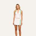 A woman stands against a plain light background wearing the Monica Tennis Polo, a sleeveless white top made from breathable fabric with green trim, paired with a matching white tennis skirt featuring green stripes.
