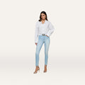 A woman stands facing forward against a plain white background, wearing a white button-up shirt, light blue Monique Skinny Jean crafted from stretch denim, a black belt, and beige high-heeled sandals.