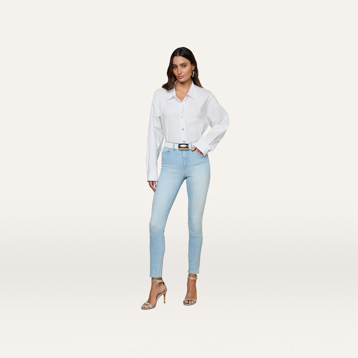A woman stands facing forward against a plain white background, wearing a white button-up shirt, light blue Monique Skinny Jean crafted from stretch denim, a black belt, and beige high-heeled sandals.