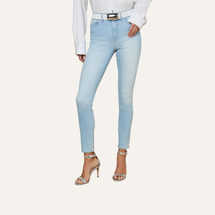 A person wearing the Monique Skinny Jean in light blue high rise stretch denim, paired with a white shirt, a white belt, and beige high-heeled sandals, is shown from the shoulders down against a plain background.