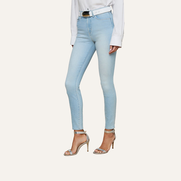 A woman wearing the Monique Skinny Jean in light blue stretch denim, paired with a white long-sleeve shirt, a white belt, and silver high-heeled sandals stands against a plain light background.