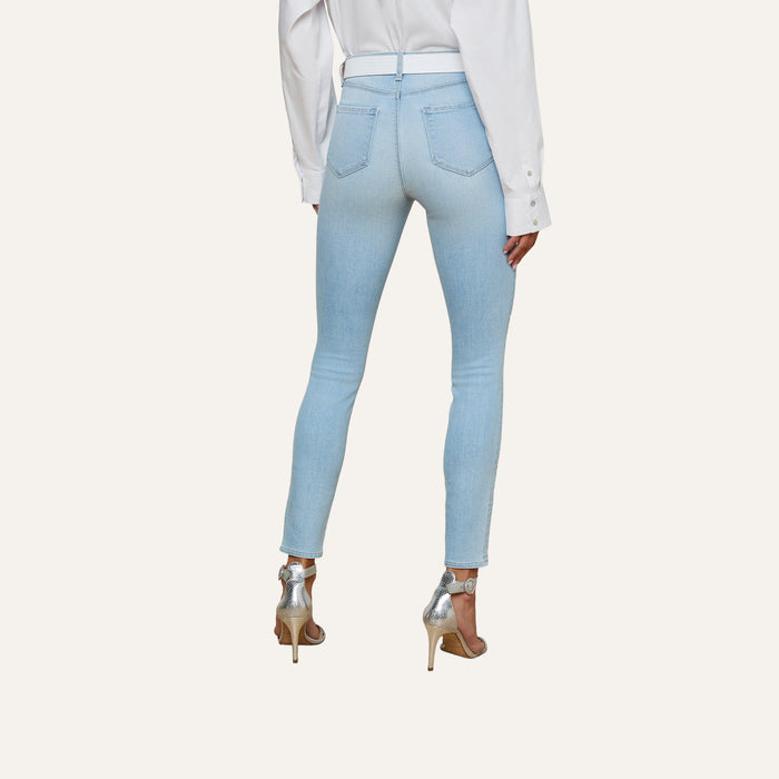 Wearing the Monique Skinny Jean in light blue stretch denim, paired with a white long-sleeve shirt and silver high-heeled sandals, a person stands facing away against a plain light background.