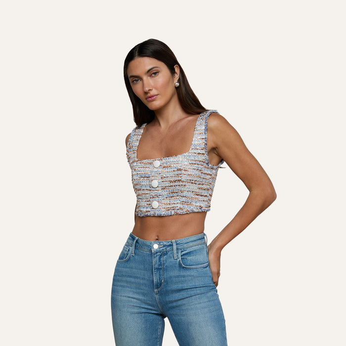 A woman stands with one hand on her hip against a plain beige background, wearing the Muno Tweed Top—a sleeveless, textured cropped top with large buttons—paired with high-waisted blue jeans.