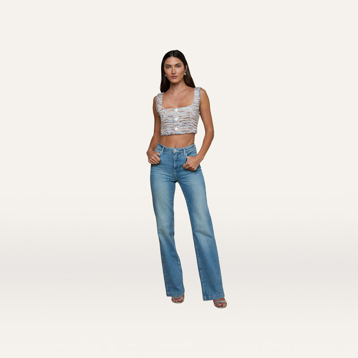 A woman stands against a plain background wearing blue jeans, heeled sandals, and the Muno Tweed Top—a textured, sleeveless cropped top with frayed edges. She has long dark hair and her hands are in her pockets.