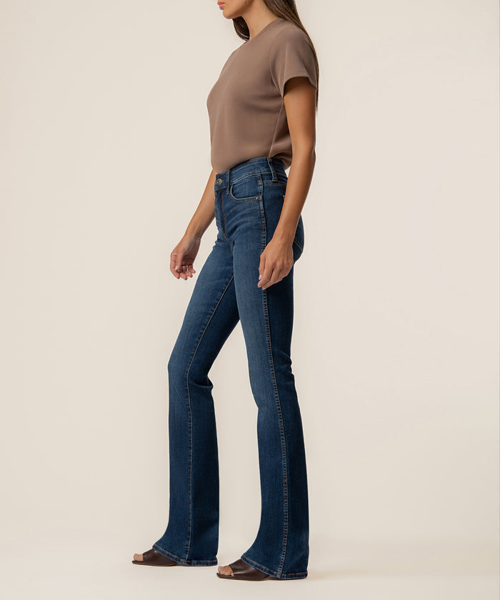 Woman wearing high-rise bootcut jeans in dark blue denim with flared hem, paired with taupe short-sleeve shirt.