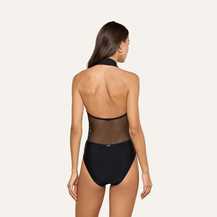 A woman with long brown hair models the Natalya Collared One-Piece Swimsuit, a black mesh halter-neck design with a sheer back panel, standing against a plain light background and facing away from the camera.