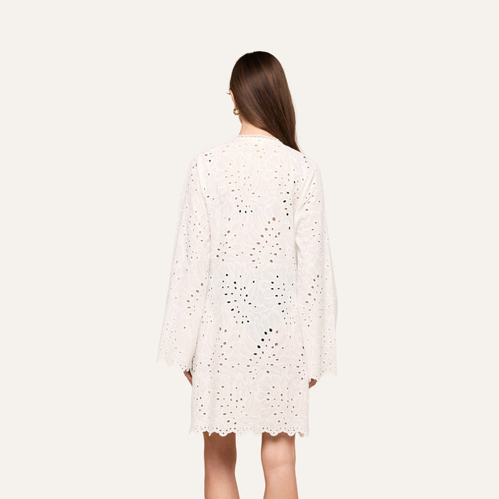 A woman with long brown hair, seen from behind, wears the Navaeh Cover-Up Kimono—a white, long-sleeve, knee-length cotton eyelet dress featuring a scalloped hem—against a plain light background.