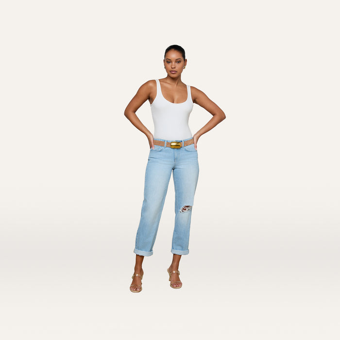 A woman stands with hands on hips, wearing a white tank top and the Nevia Slouchy Straight-Leg Jean in a light wash with rolled cuffs, paired with a belt and heeled sandals against a plain, light background.
