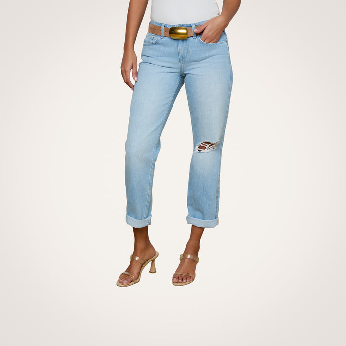 The woman is wearing Nevia Slouchy Straight-Leg Jeans in a light wash with a ripped knee, paired with a brown belt, tan heeled sandals, and a white top. Her right hand is in her pocket against a plain white background.