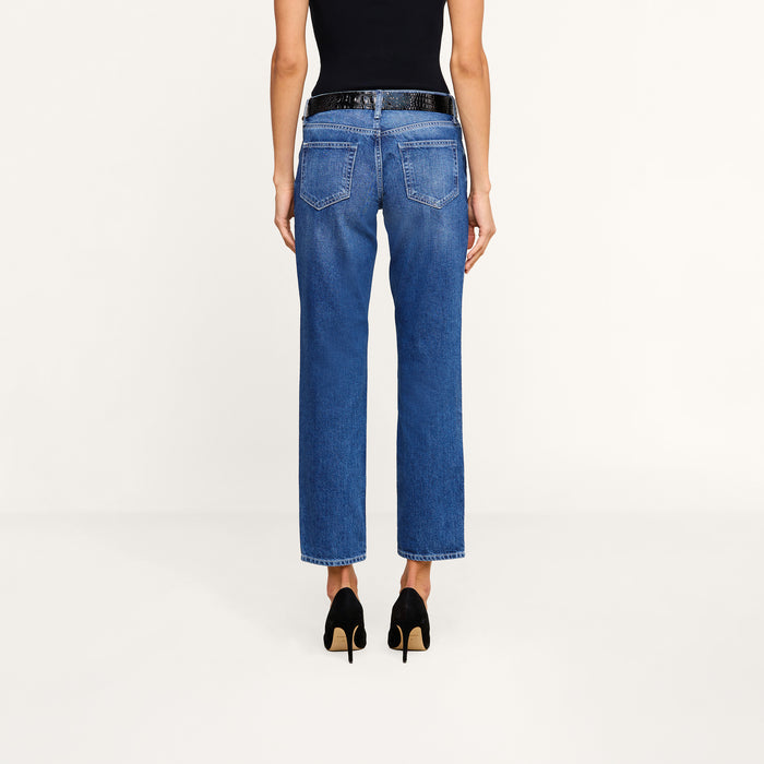 A person stands facing away, wearing a black sleeveless top, the Nevia Slouchy Straight-Leg Jean in vintage-inspired blue high-waisted all-cotton denim, and black high-heeled shoes against a plain white background.
