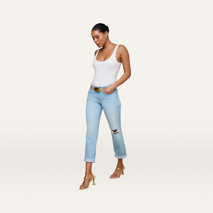 A woman stands against a plain white background, looking down with one hand in her pocket, wearing a white tank top, tan heeled sandals, a brown belt, and the Nevia Slouchy Straight-Leg Jean in light wash denim with rips.