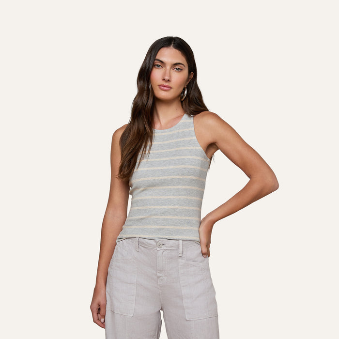 A woman wearing the Nia Ribbed Tank—a sleeveless, light gray and white striped racerback top—paired with light-colored pants stands against a plain background with one arm resting on her hip.