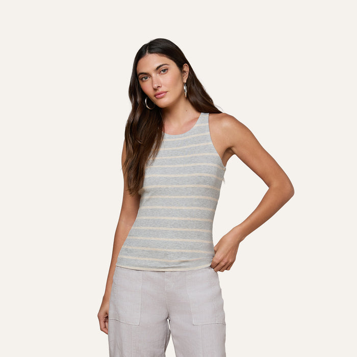 A woman with long brown hair is wearing the Nia Ribbed Tank, a sleeveless gray and white striped top made from soft microfiber modal, paired with light-colored pants and standing against a plain light background.