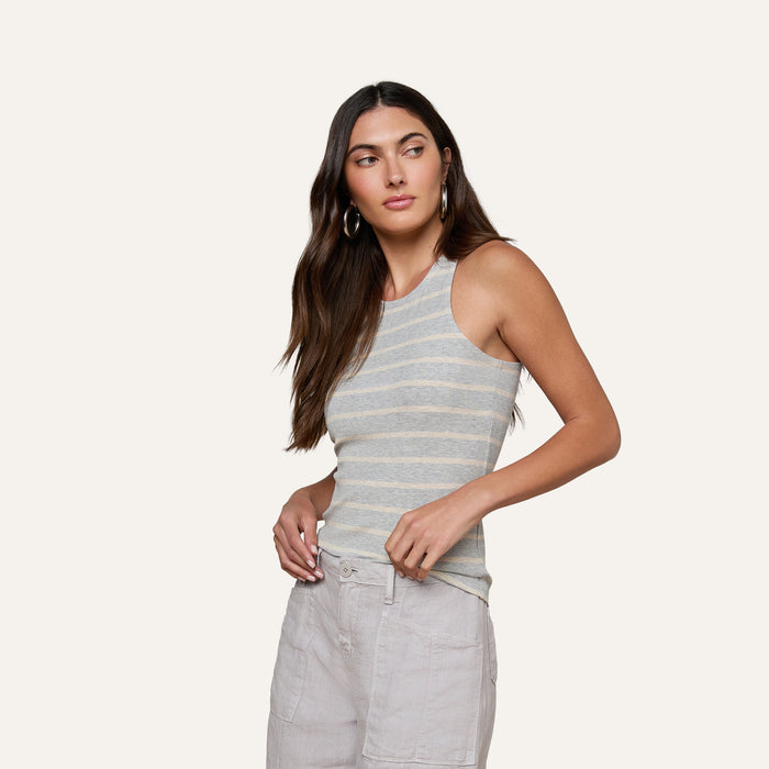 A woman with long brown hair, wearing hoop earrings, a sleeveless gray and white striped Nia Ribbed Tank and light-colored pants, stands against a plain light background, looking slightly to her left.