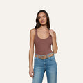 A woman with long brown hair stands against a plain background, wearing the Nica Scoopneck Bodysuit in brown, paired with light blue jeans and a tan belt. She is holding one strap of her top with her right hand.