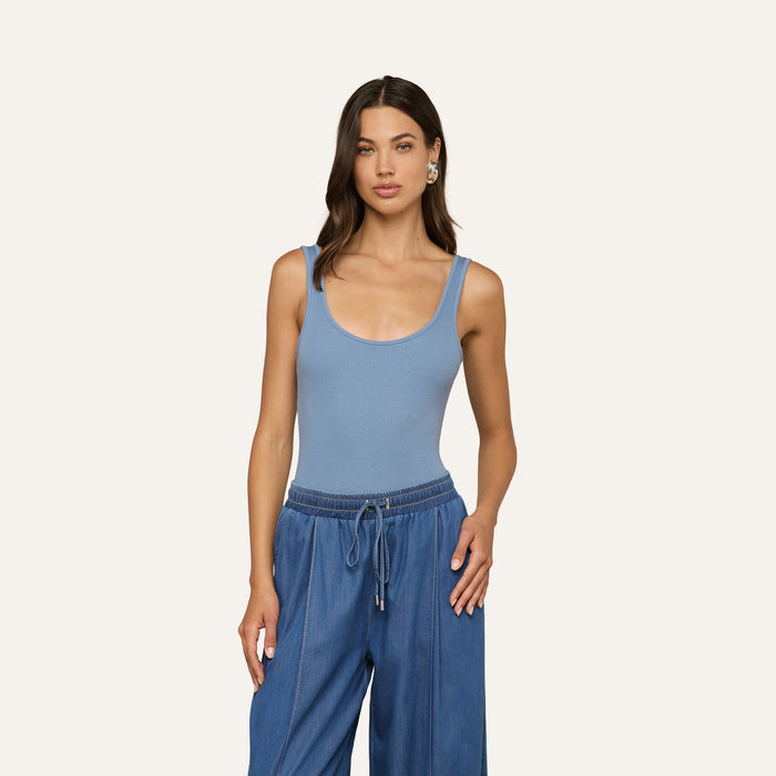 A woman wearing a light blue Nica Scoopneck Bodysuit and loose blue drawstring pants stands against a plain light background, looking at the camera with a neutral expression.
