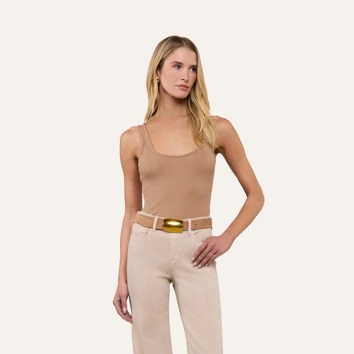 Woman with long blonde hair wearing the Nica Scoopneck Bodysuit in tan, paired with beige pants and a wide belt with a gold buckle, standing against a plain light background.