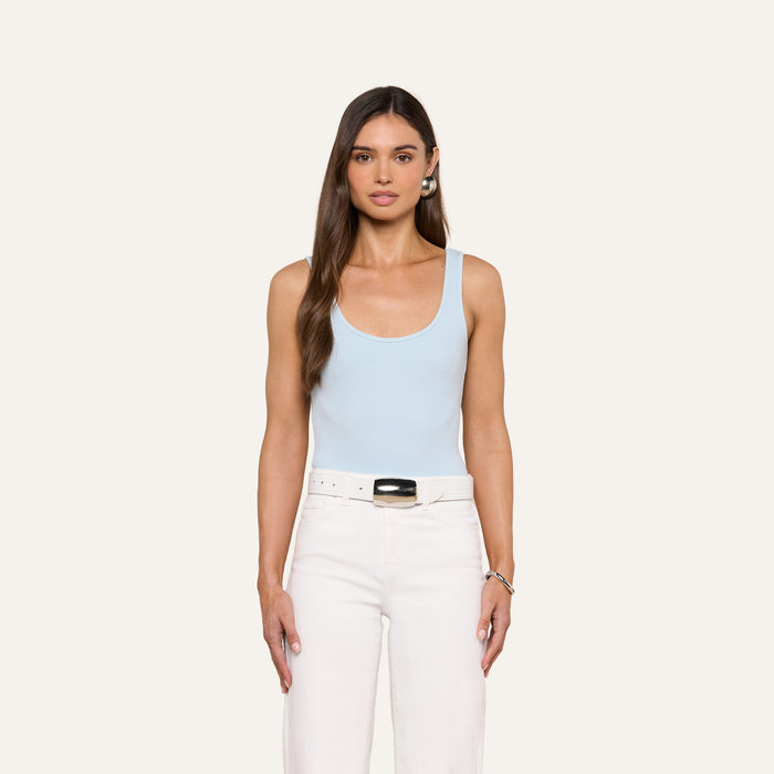 A woman with long brown hair and a neutral expression stands against a plain light background, wearing the Nica Scoopneck Bodysuit in light blue, styled with white pants, a white belt, and a bracelet. Her arms are relaxed at her sides.