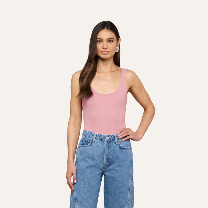 A woman with long brown hair wears the Nica Scoopneck Bodysuit and high-waisted blue jeans, standing with one hand on her hip against a plain light background.