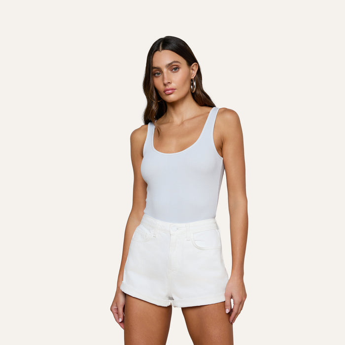A woman with long brown hair wears the Nica Scoopneck Bodysuit paired with high-waisted white shorts, standing against a plain light background.