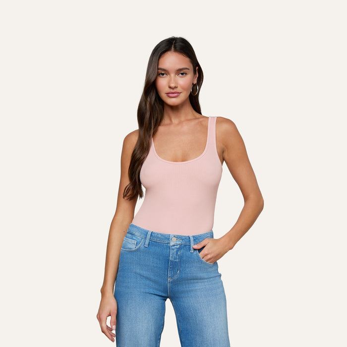 A woman with long brown hair stands against a plain off-white background, looking at the camera with a neutral expression while wearing the Nica Scoopneck Bodysuit in light pink and blue jeans.