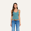 The woman stands facing forward against a plain light background, wearing the Nica Scoopneck Bodysuit in teal paired with blue jeans and a brown belt. The bodysuit’s sculpted fit and high-recovery stretch rib highlight her silhouette. She has long, dark hair and minimal accessories.