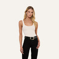 A woman with long blonde hair wears the Nica Scoopneck Bodysuit, a white stretch-cotton piece featuring a scoop neckline. She pairs it with black pants and a black belt with a large buckle, standing facing forward against a plain light background.