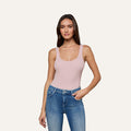 A woman with long brown hair stands against a plain light background, looking at the camera with a slight smile while wearing the Nica Scoopneck Bodysuit in light pink and blue jeans.