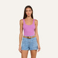 A woman stands against a plain light background wearing a sleeveless pink Nica Scoopneck Bodysuit made of high recovery stretch rib, paired with light blue denim shorts featuring large front pockets and a brown belt.
