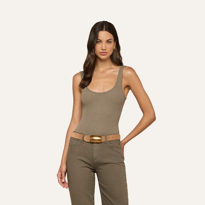Woman with long, wavy brown hair wearing a sculpted fit sleeveless smoky olive green Nica Scoopneck Bodysuit, matching olive pants, and a tan belt, standing with one hand on her hip against a plain light background.