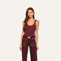 A woman with long brown hair wears a sleeveless maroon Nica Scoopneck Bodysuit paired with matching leather pants and a wide belt, standing against a plain light background, looking forward with her arms relaxed at her sides.