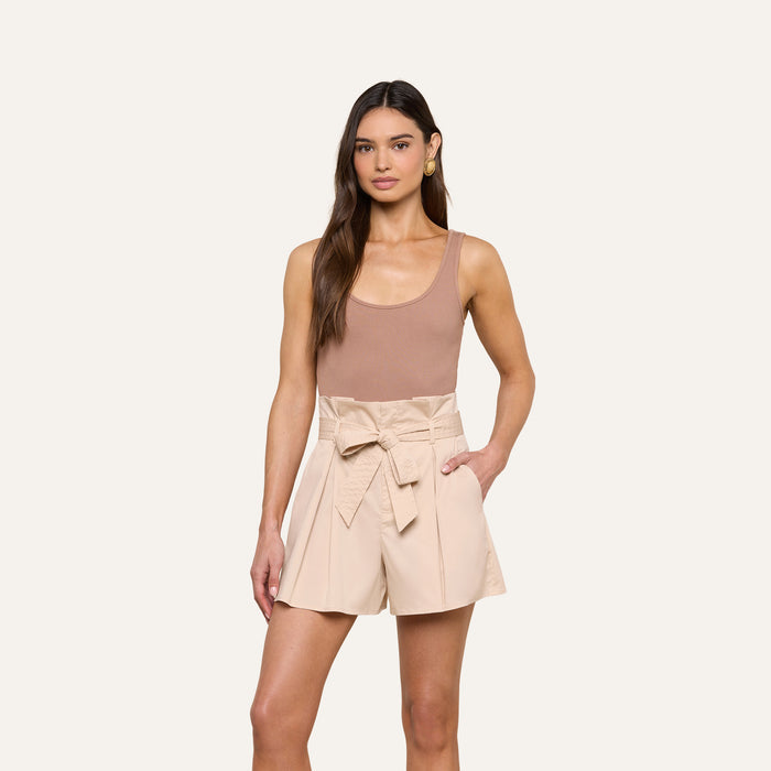 A woman stands against a plain background wearing the Nica Scoopneck Bodysuit—a sleeveless tan top with a sculpted fit—paired with high-waisted, light beige shorts featuring a tied belt, one hand in her pocket.