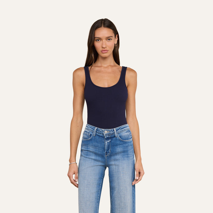A woman with long brown hair wears the Nica Scoopneck Bodysuit in navy blue, styled with light blue high-waisted jeans, standing against a plain off-white background.