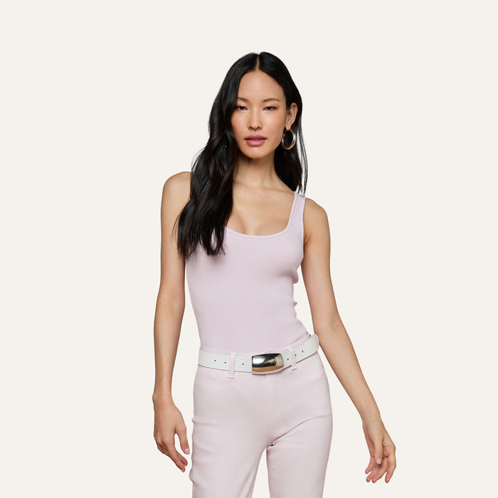 A woman in the Nica Scoopneck Bodysuit in light pink, paired with matching pink pants, a white belt with a silver buckle, and hoop earrings stands against an off-white background.
