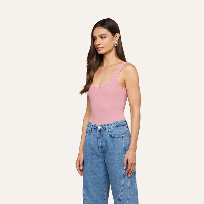 A woman with long brown hair wears the pink Nica Scoopneck Bodysuit, featuring a stretch rib texture, paired with blue high-waisted jeans and large earrings, as she stands against a plain light background looking slightly to the side.