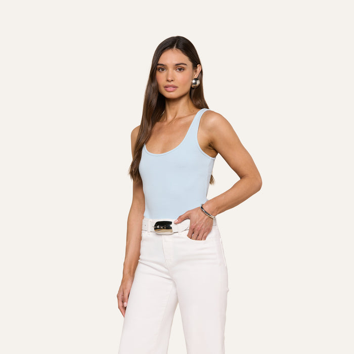 A woman with long brown hair wears the Nica Scoopneck Bodysuit in light blue and high-waisted white pants, standing against a plain off-white background with one hand in her pocket and a neutral expression.