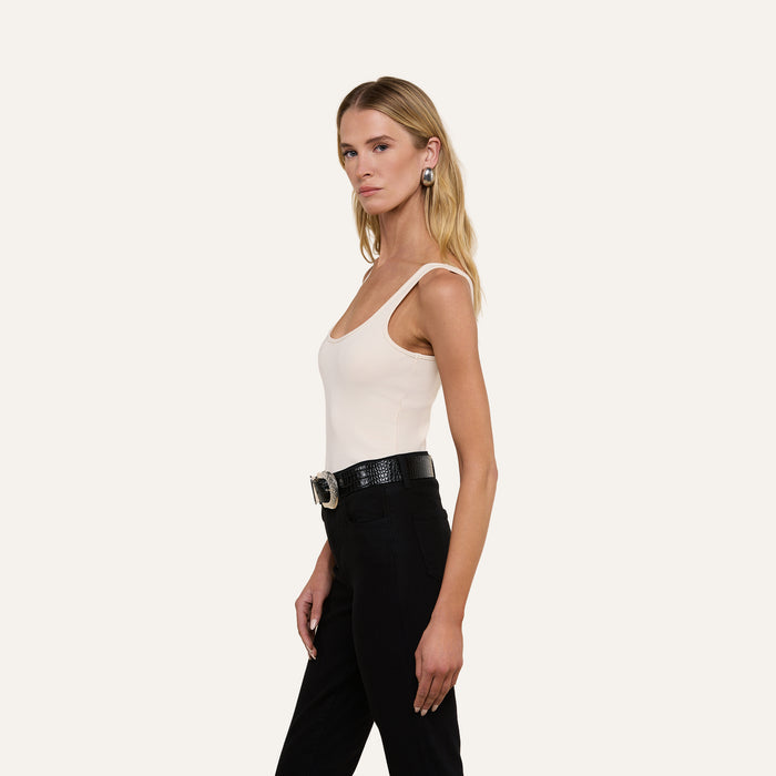 A woman with long blonde hair is wearing the Nica Scoopneck Bodysuit, styled with black pants, a black belt featuring a large buckle, and earrings. She stands against a plain light background, facing left in a relaxed pose.