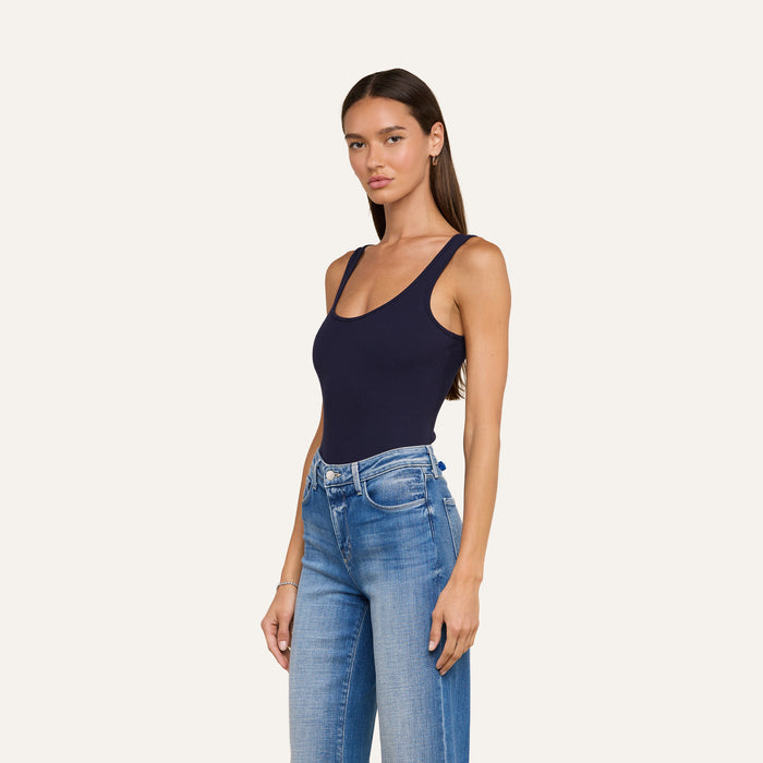 A woman with long brown hair wears the Nica Scoopneck Bodysuit in navy and high-waisted blue jeans, standing against a plain off-white background and looking at the camera with a neutral expression.