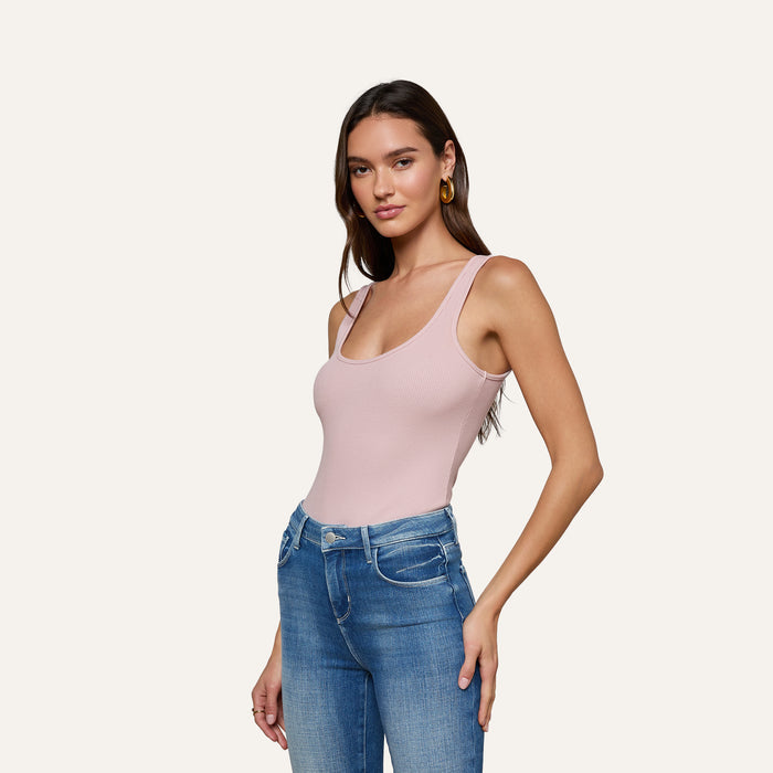 A woman wearing the Nica Scoopneck Bodysuit in light pink with blue jeans stands against a plain off-white background, posing with one hand on her hip and looking at the camera.