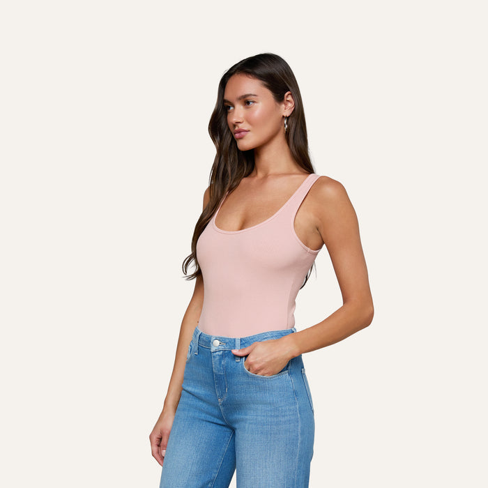 A woman with long brown hair wears the Nica Scoopneck Bodysuit in light pink paired with blue jeans, standing with one hand in her pocket and gazing to the side against a plain light background.