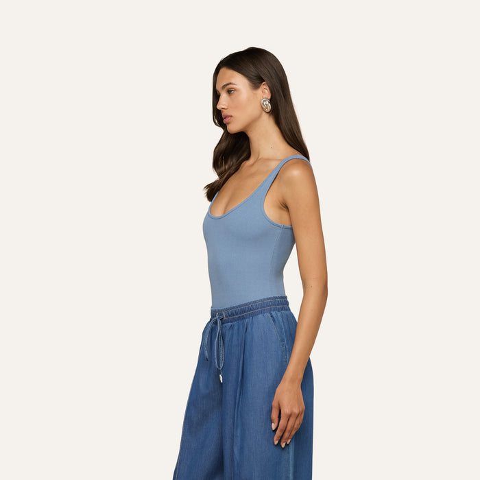 Woman with long brown hair wearing the Nica Scoopneck Bodysuit in light blue, featuring a sleeveless and sculpted fit, paired with wide-leg blue drawstring pants and large round earrings, standing in profile against a plain light background.