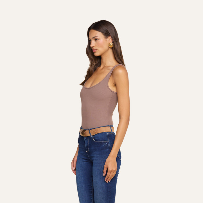 A woman stands in profile against a plain light background, wearing the Nica Scoopneck Bodysuit— a taupe stretch rib tank with a sculpted fit—paired with blue jeans featuring cutouts at the waist and gold earrings.