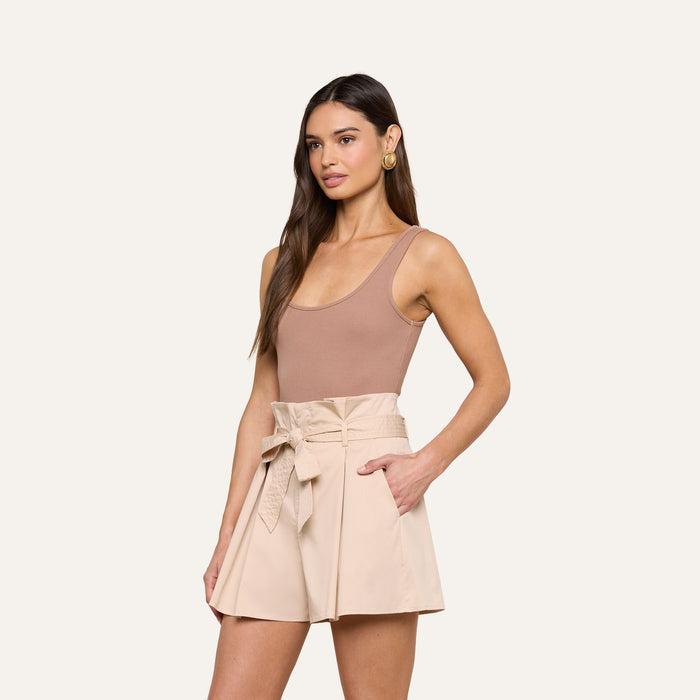 A woman wearing the Nica Scoopneck Bodysuit in sleeveless, scoop-neck beige with high recovery stretch rib and high-waisted, light beige shorts with a tied belt stands with one hand in her pocket against a plain background.