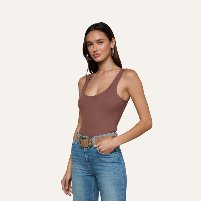 A woman stands against a plain light background, posing with one hand in her pocket, wearing the Nica Scoopneck Bodysuit in brown and blue jeans with a tan belt.